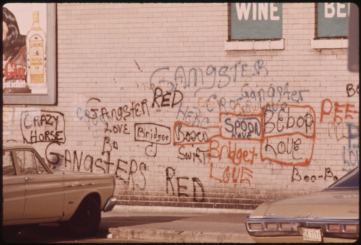 A pink brick wall in an urban street covered with layered graffiti in black, blue, and orange marker and paint, with handwritten text and names.