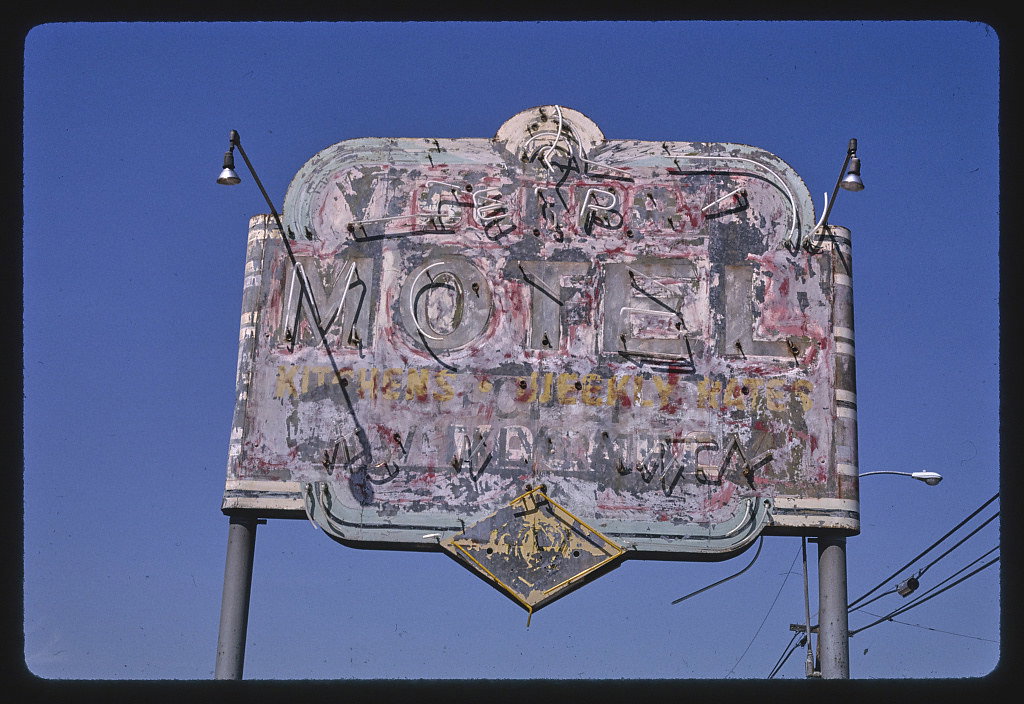 A weathered vintage motel sign photographed against a clear blue sky, featuring large dimensional lettering with ornate neon tubing and flanking lamp fixtures.