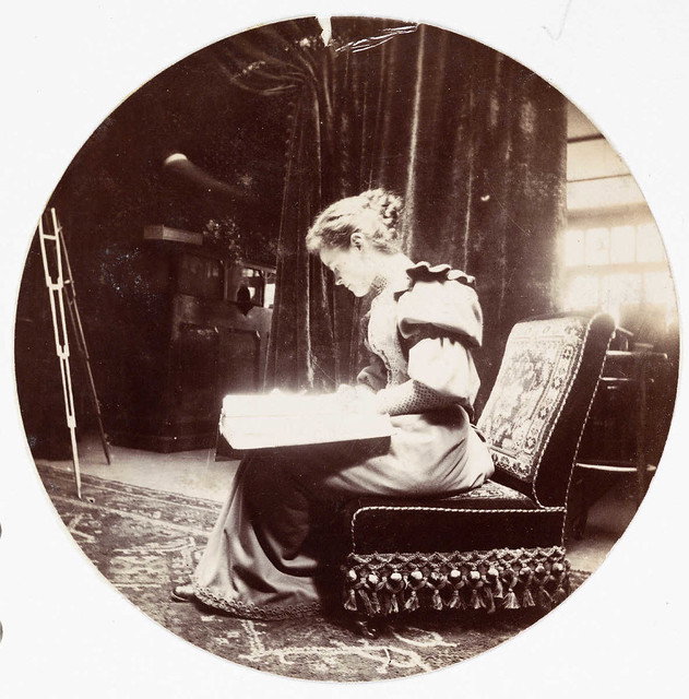 A sepia-toned photograph from 1890 showing a woman seated indoors reading a book, wearing period dress with puffed sleeves.