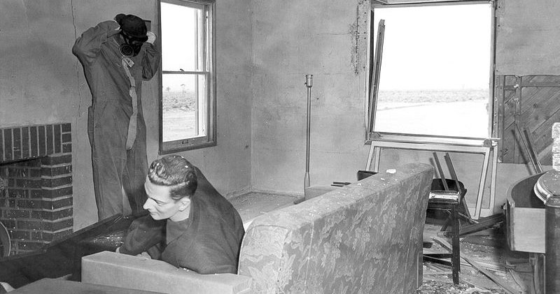 ‘Damaged living room with mannequins. One inspector observing area’ by National Nuclear Security Administration (1953)